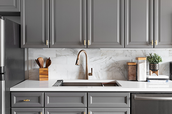 A kitchen sink detail shot with grey cabinets, a white marble co